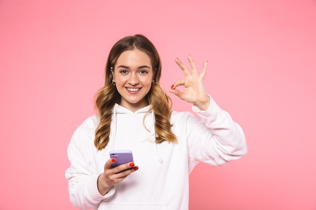 Sonriente mujer rubia vestida con ropa casual que muestra el signo de ok y mirando al frente mientras sostiene el teléfono inteligente sobre la pared rosa