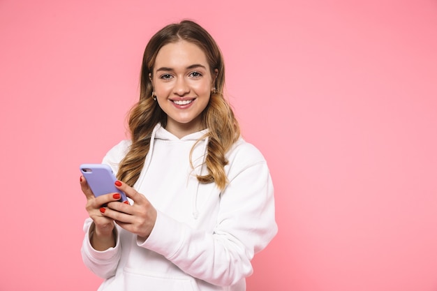 Sonriente mujer rubia vestida con ropa casual sosteniendo el teléfono inteligente y mirando al frente sobre la pared rosa