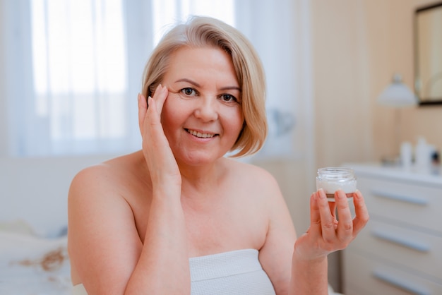 Sonriente mujer senior aplicando loción antienvejecimiento para eliminar las ojeras bajo los ojos.