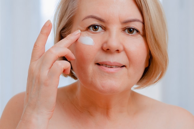 Sonriente mujer senior aplicando loción antienvejecimiento para eliminar las ojeras bajo los ojos.