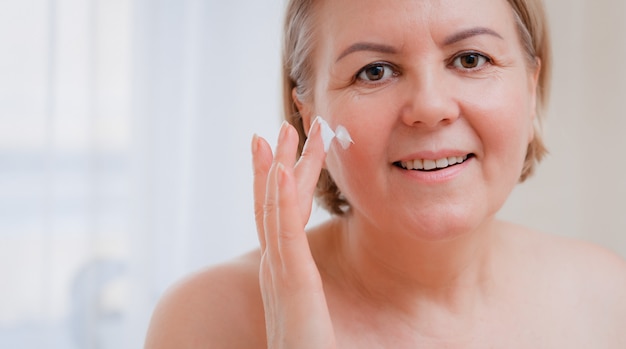 Foto sonriente mujer senior aplicando loción antienvejecimiento para eliminar las ojeras bajo los ojos.