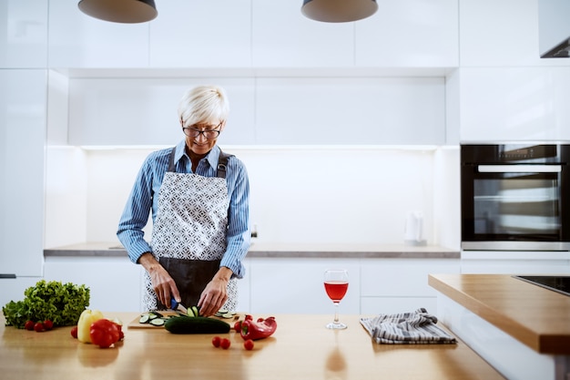 Sonriente mujer senior en delantal de pie en la cocina y cortar pepino