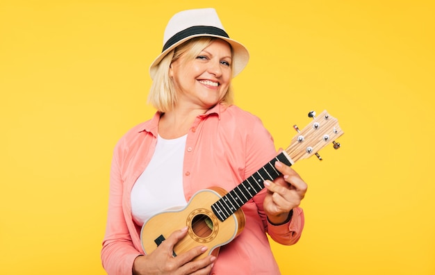 Sonriente mujer senior rubia feliz linda de buen humor está posando con ukelele sobre fondo amarillo