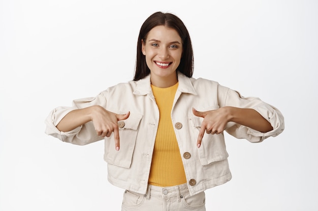 Sonriente mujer sincera, agradable expresión de rostro amistoso, señalando con el dedo hacia abajo, mostrando el anuncio de venta debajo, de pie en blanco