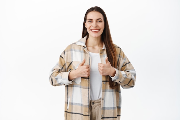 Sonriente mujer solidaria con una hermosa sonrisa, muestra los pulgares hacia arriba en señal de aprobación, le gusta el producto o la marca, apoya, elogia la calidad del trabajo realizado, de pie sobre una pared blanca