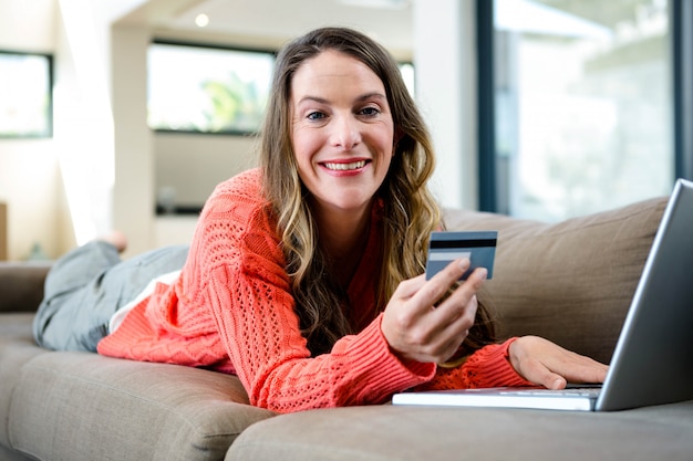 sonriente mujer tendida en el sofá con su computadora portátil y tarjeta de crédito