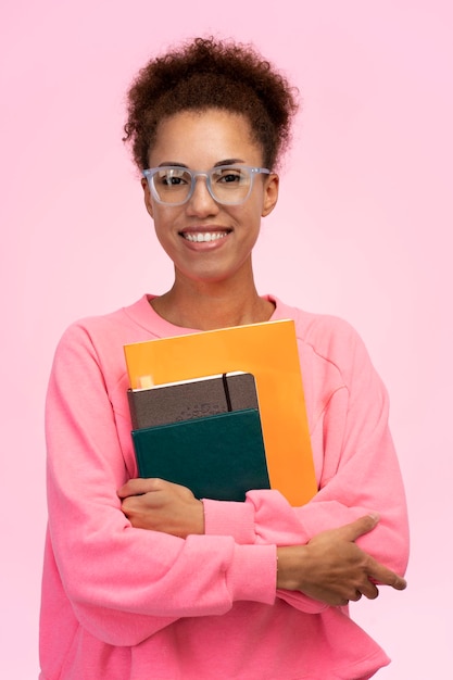 Sonriente niña adolescente africana estudiante de secundaria con gafas posando con libros sobre fondo rosa