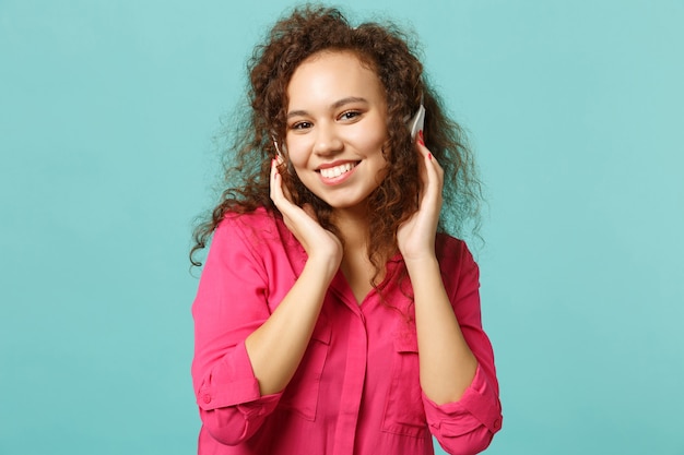 Sonriente niña africana en ropa casual mirando a cámara, escuchando música con auriculares aislados sobre fondo azul turquesa en estudio. Personas sinceras emociones, concepto de estilo de vida. Simulacros de espacio de copia.