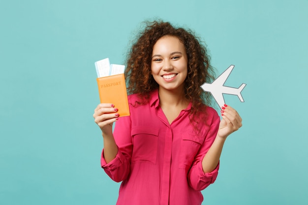 Sonriente niña africana en ropa casual con pasaporte, boleto de embarque, avión de papel aislado sobre fondo de pared azul turquesa. Concepto de estilo de vida de emociones sinceras de personas. Simulacros de espacio de copia.