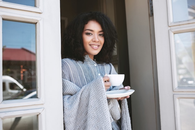 Sonriente niña afroamericana apoyado en la puerta con una taza de café envuelto en cuadros. Bella dama con cabello rizado oscuro tomando café en el restaurante