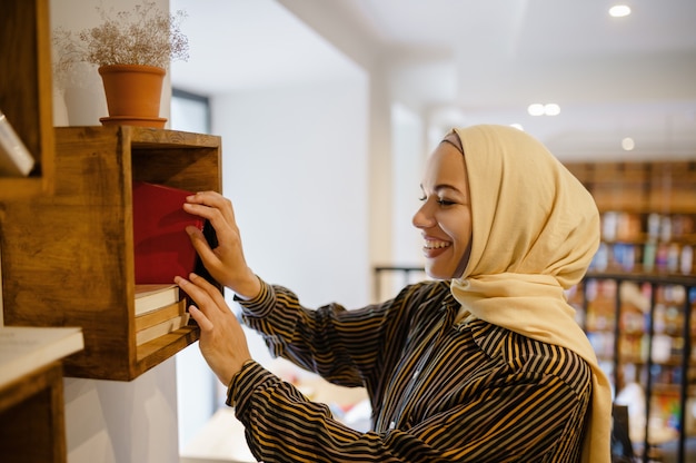 Sonriente niña árabe en hijab en la estantería, interior del café de la universidad en el fondo. Mujer musulmana con libros sentado en la biblioteca.