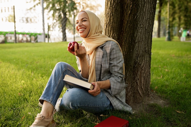 Sonriente niña árabe en hijab tiene manzana y libros de texto en el parque de verano. Mujer musulmana con libros descansando sobre el césped.