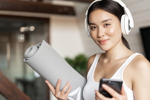 Sonriente niña asiática en auriculares escuchando música en casa y entrenamiento con tapete sosteniendo teléfono móvil ...