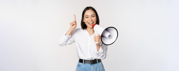 Sonriente niña asiática feliz hablando en megáfono y apuntando hacia arriba anunciando promoción de descuento mostrando adve