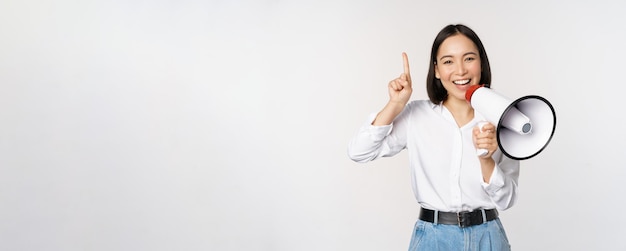 Sonriente niña asiática feliz hablando en megáfono y apuntando hacia arriba anunciando promoción de descuento mostrando adve