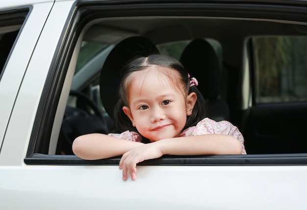 Sonriente niña asiática mirando la cámara desde la ventana del coche blanco.