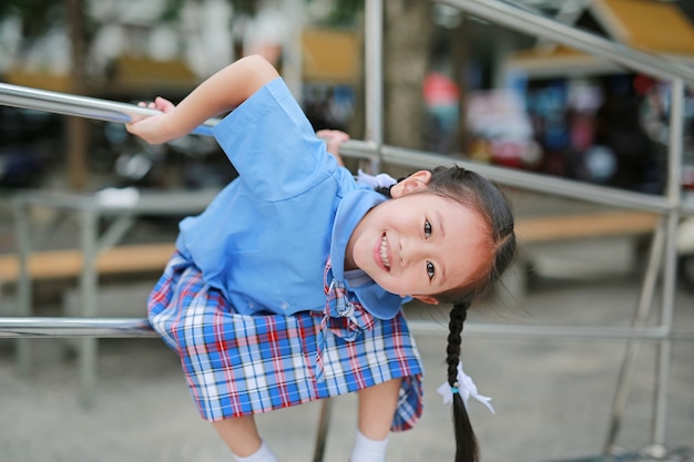 Sonriente niña asiática en uniforme escolar colgando en la barandilla de metal.