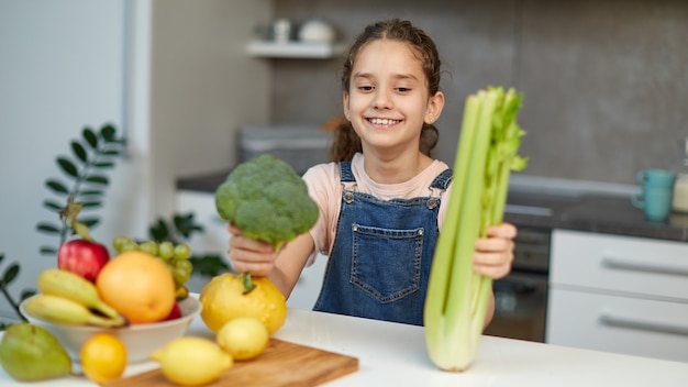 Sonriente niña bonita de pie junto a la mesa en la cocina, sosteniendo brócoli verde y apio en las manos.