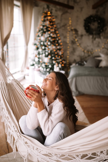 Sonriente niña de cabello oscuro vestida con suéter beige y pantalones sostiene una taza roja sentada en una hamaca en una acogedora habitación decorada con un árbol de año nuevo. .