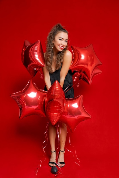 Sonriente niña caucásica feliz en vestido negro con globos rojos en forma de estrellas