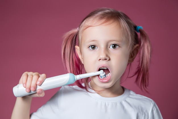 Sonriente niña caucásica limpiando sus dientes con cepillo de dientes sónico eléctrico sobre fondo rosa.