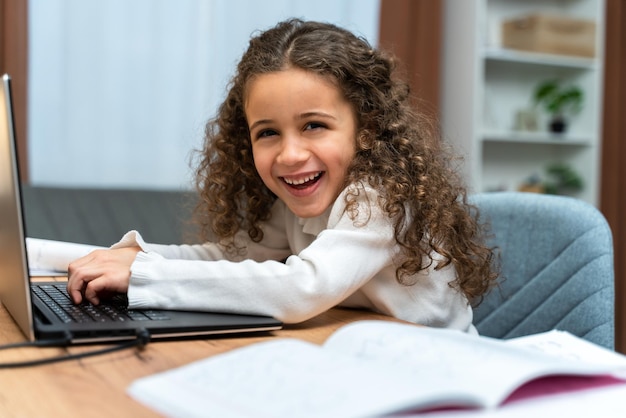 Sonriente niña caucásica riendo a carcajadas y mirando a la cámara mientras usa la computadora portátil Estudiar en línea sobre el concepto de educación en el hogar