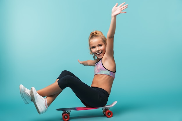 Sonriente niña de deportes jugando con una patineta aislada sobre pared azul