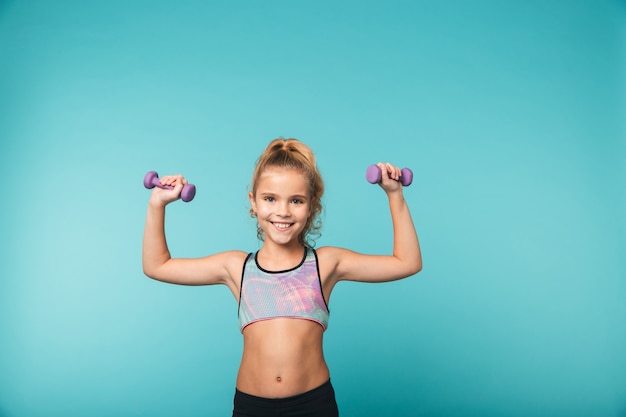 Sonriente niña deportiva haciendo ejercicios con pesas aislado sobre pared azul