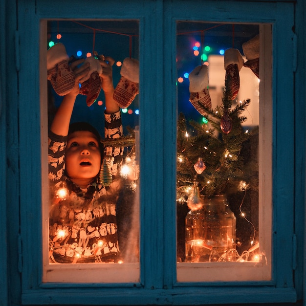 Sonriente niña dulce niño ventana permanente tiempo de Navidad. Con luces de colores Fondo de árbol de Navidad, enfoque selectivo. Anticipación vacaciones, sueños, regalos, noche de año nuevo 2022, estilo rústico retro