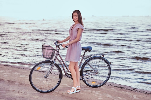 Sonriente niña encantadora con vestido camina con su bicicleta en la playa.