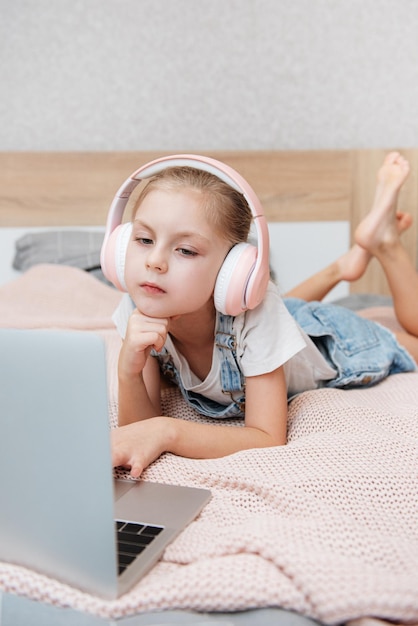 Sonriente niña feliz usando laptop en auriculares inalámbricos en la cama en casa.