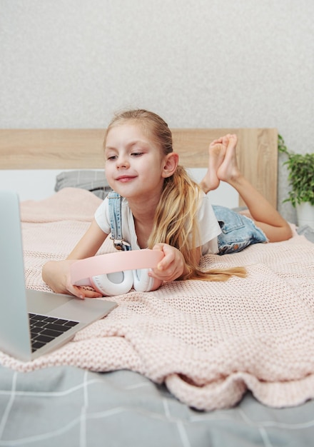 Sonriente niña feliz usando laptop en auriculares inalámbricos en la cama en casa.