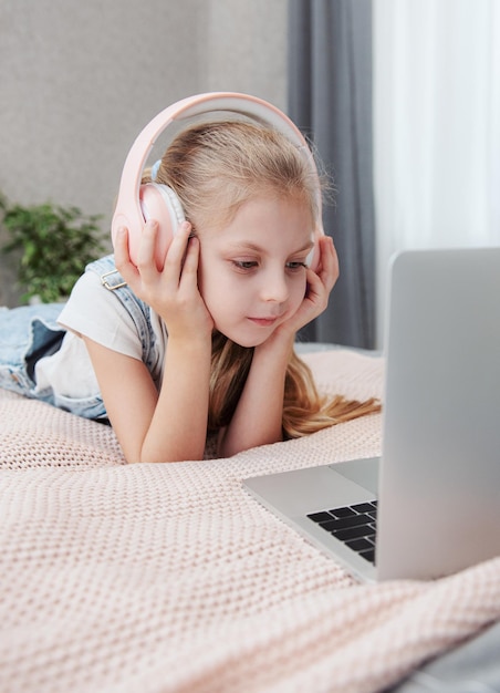 Sonriente niña feliz usando laptop en auriculares inalámbricos en la cama en casa