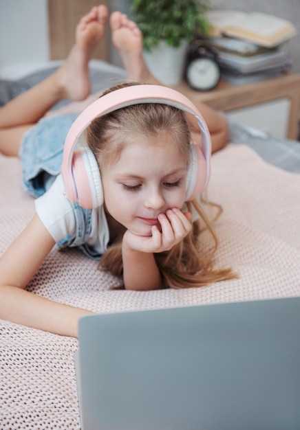 Sonriente niña feliz usando laptop en auriculares inalámbricos en la cama en casa