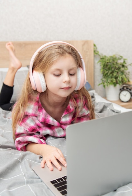 Sonriente niña feliz usando laptop en auriculares inalámbricos en la cama en casa