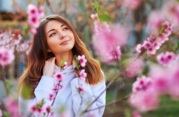 Sonriente niña hermosa de pie cerca de un melocotonero durante la puesta de sol. Cara feliz. Tiempo de primavera.