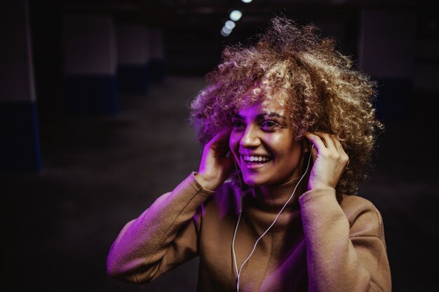 Sonriente niña de hip hop de raza mixta de pie en el garaje subterráneo y disfrutando de la música.