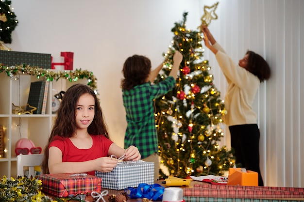 Sonriente niña hispana envolver regalos cuando sus amigos decoran el árbol de Navidad en segundo plano.