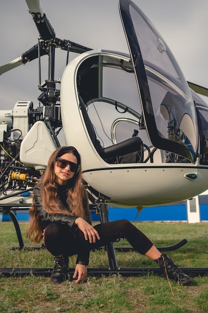 Sonriente niña de interpolación sentada cerca de helicóptero abierto en campo de vuelo