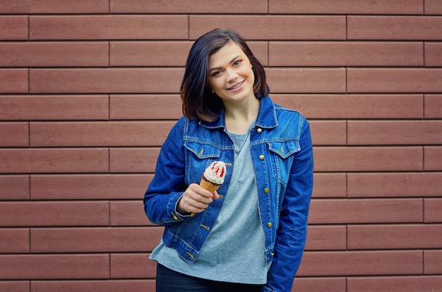 Sonriente niña morena caucásica sosteniendo en una mano sabroso helado con mermelada de frutos rojos en una chaqueta de jeans cerca de una pared de ladrillo con textura marrón.