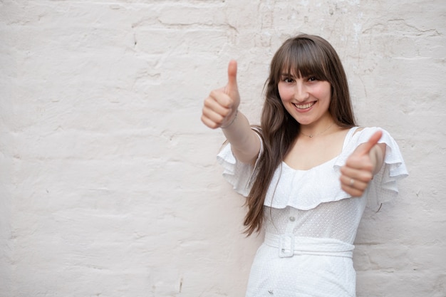 Sonriente niña morena con el pelo largo con un vestido blanco muestra un signo de clase con un pulgar levantado sobre un fondo de ladrillo macizo. lugar para tu diseño