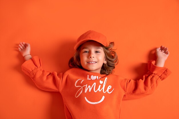 Foto sonriente niña niño 6-7 años en gorra y ropa de moda se encuentra en el piso en el estudio sobre fondo naranja. copia espacio