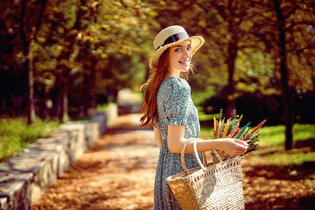 Sonriente niña pelirroja camina sola en el parque de otoño en un día cálido y soleado mientras sostiene el bolso de verano indio