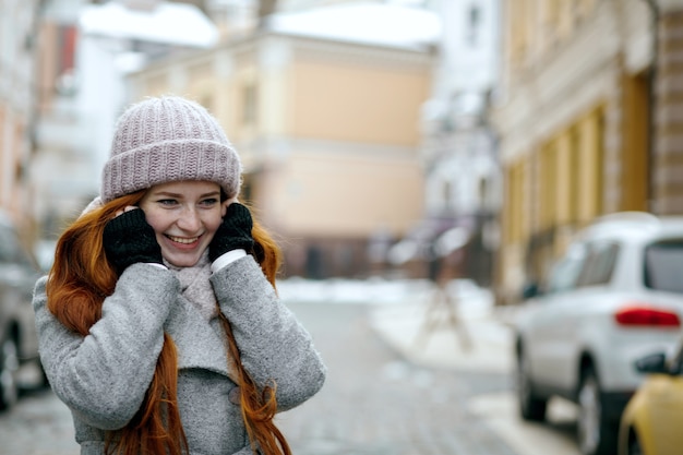 Sonriente niña de pelo rojo con ropa de abrigo de invierno caminando por la calle. Espacio para texto