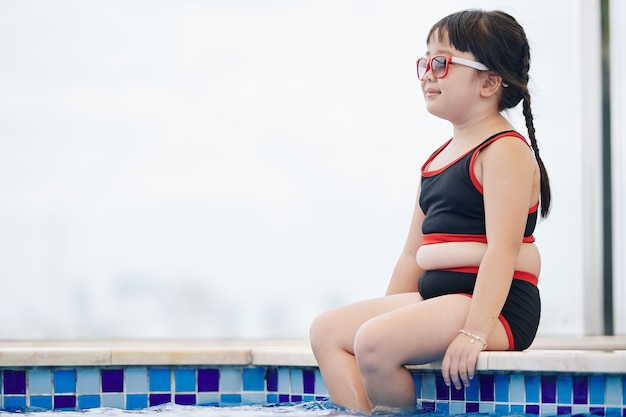 Sonriente niña preadolescente en traje de baño y gafas de sol sentado en el borde de la piscina