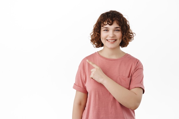 Sonriente niña sincera con cabello rizado que señala el dedo a la izquierda, mostrando copia espacio de pie en camiseta en blanco