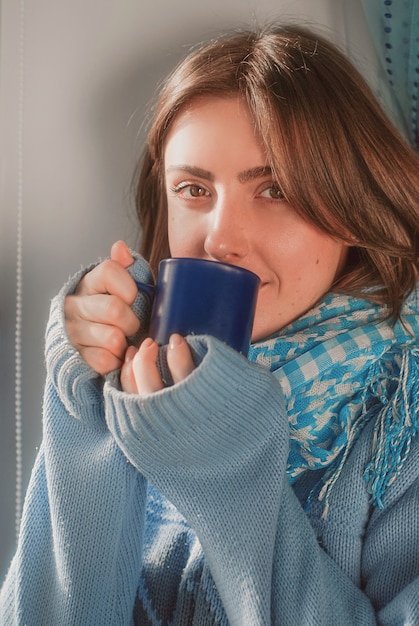 Foto sonriente niña soñadora en suéter con la taza cerca de la ventana congelada en el invierno