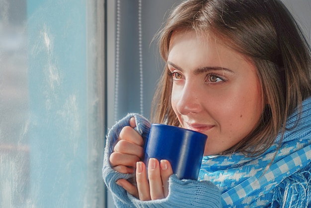 Sonriente niña soñadora en suéter con la taza cerca de la ventana congelada en el invierno
