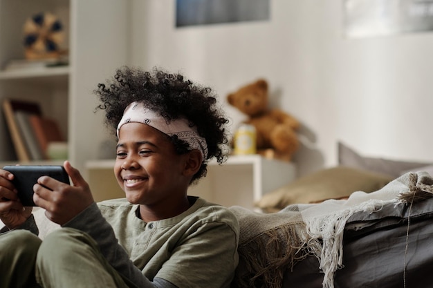 Sonriente niño afroamericano en diadema mirando la pantalla del teléfono inteligente