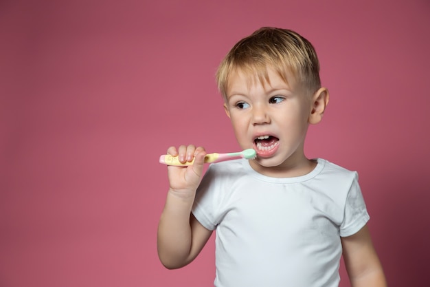 Sonriente niño caucásico limpiando sus dientes con cepillo de dientes manual para niños sobre fondo rosa.